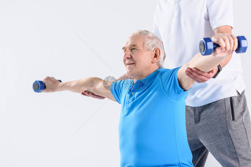 协助老年男子在灰色背景下用哑铃进行锻炼的康复治疗师的部图片