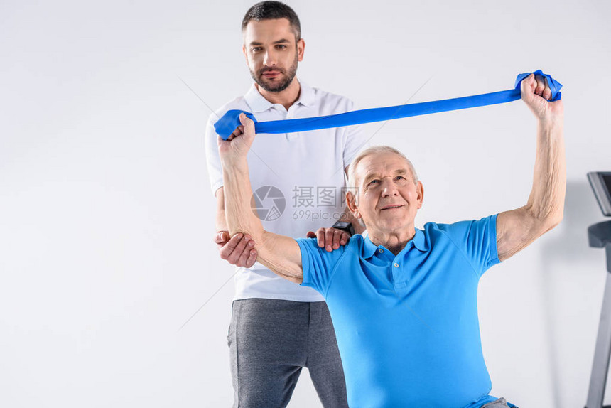 协助老年男子在灰色背景上用橡胶带进行锻炼的康图片