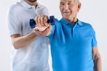 协助老年男子使用孤立在灰色地带的哑铃进行锻炼的康复治疗图片