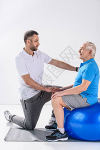 协助老年男子在灰色背景下健身球上运动的康复治疗师图片