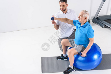 康复治疗师协助老年男子在健身球上用哑铃进行运动的康复治疗师图片