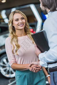 美发店助理和微笑的女顾客在汽车沙龙中握着手进行握手图片