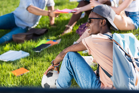 微笑的年轻非洲裔美国学生拿着足球坐在草地上背景图片