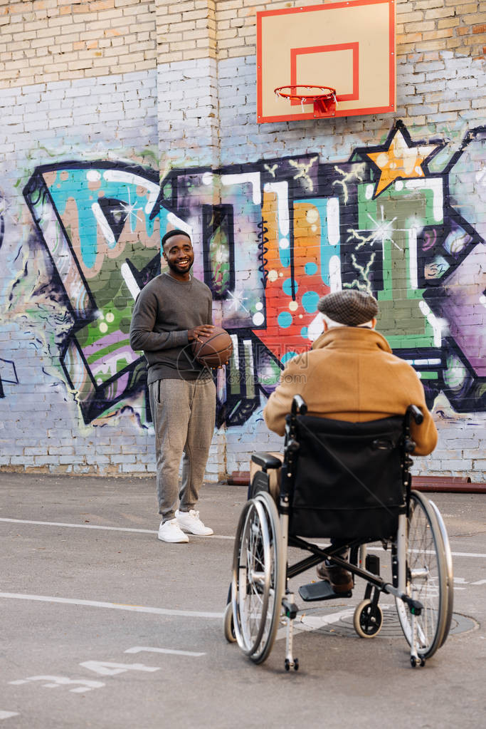 坐在轮椅上的老年残疾男子的背面观望着在街上打篮球的非图片
