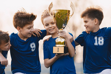 男孩运动队庆祝胜利快乐的孩子们拿着金奖杯儿童足球队举起优胜者杯青背景图片