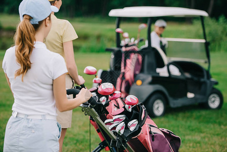 有高尔夫设备的女高尔夫球手和朋友在绿色草坪后面的高尔夫球车中图片