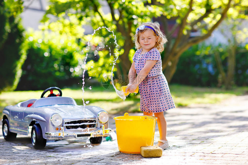 可爱的漂亮蹒跚学步的女孩在夏日花园里洗大旧玩具车图片