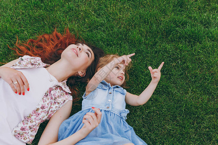 穿着轻薄连衣裙的温柔女人和小可爱的小女孩用食指向上躺在公园的绿草上图片