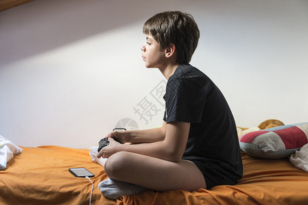 年轻男孩在充电时坐在她房间里的床上玩电子游图片