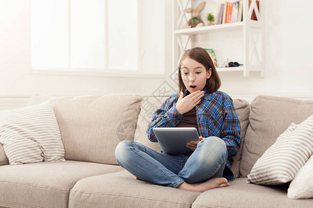 使用数字平板电脑躺在米地沙发上复制空间的年轻受惊女孩令人震惊的新闻图片