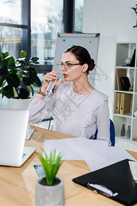 戴眼镜坐在现代办公室办公桌边和喝水杯的年轻图片