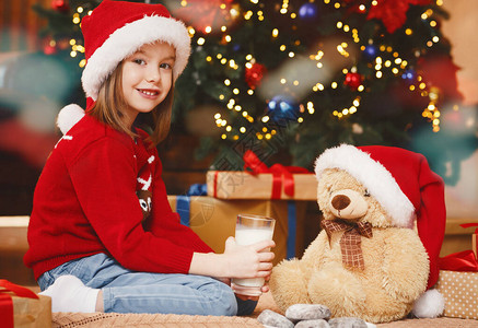 在圣诞树旁边用饼干和牛奶给泰迪熊吃饼干和奶汁的圣塔帽可爱图片