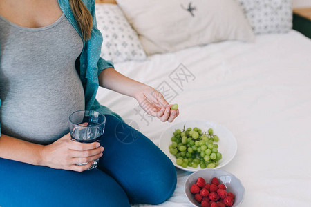 年轻孕妇用水杯在床上吃水果的照片以作图片