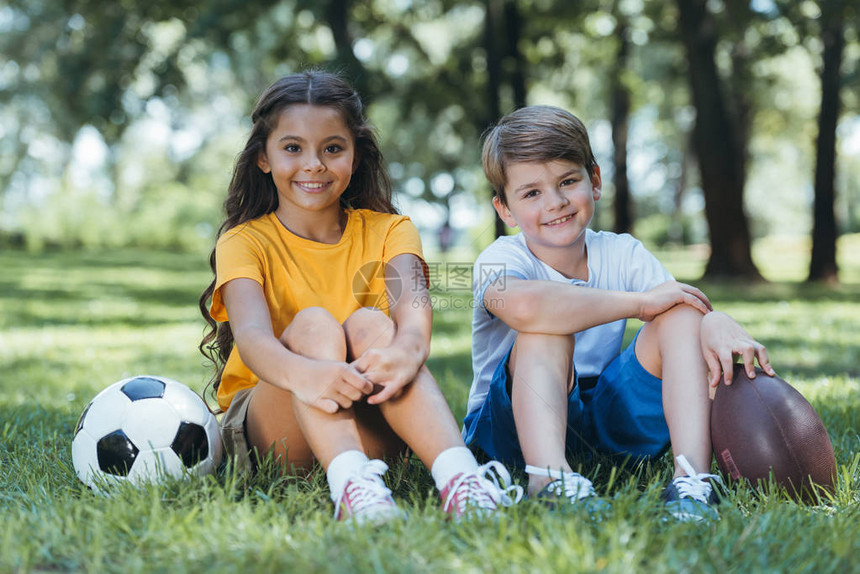 带着足球和橄榄球的可爱快乐儿童坐在公园里图片