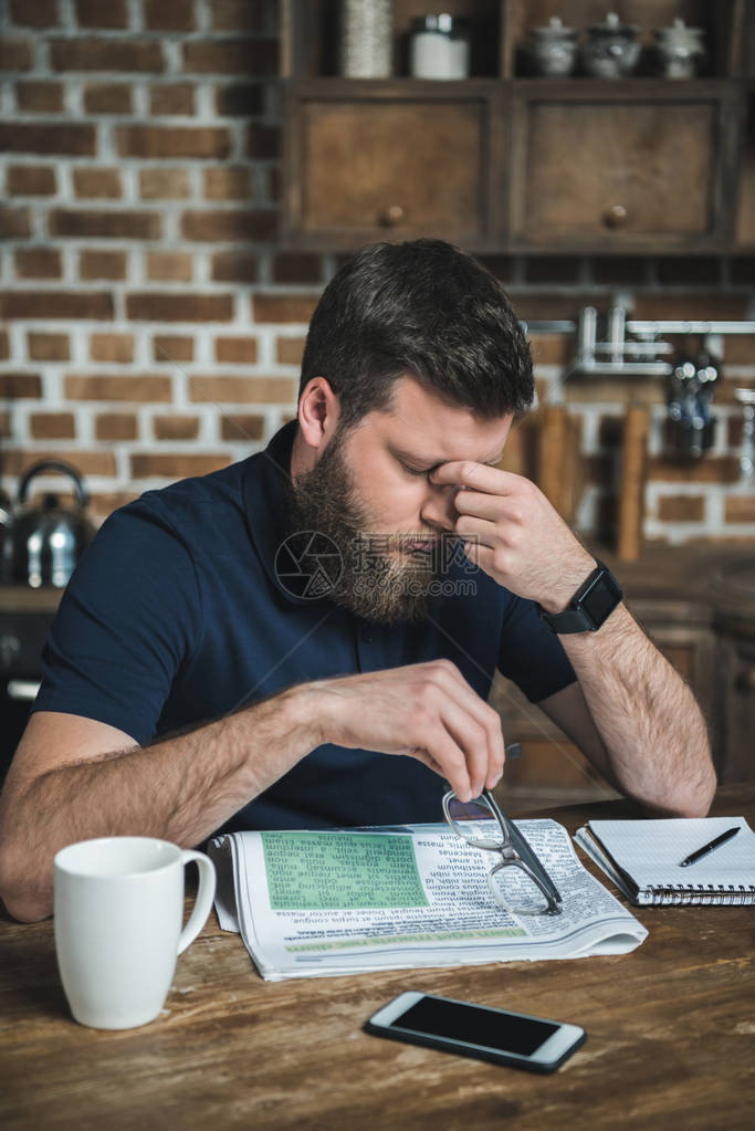 疲惫的男人在家看报时揉眼睛的画像图片