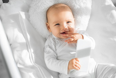 在白色婴儿床上拿着智能手机的快乐婴儿图片