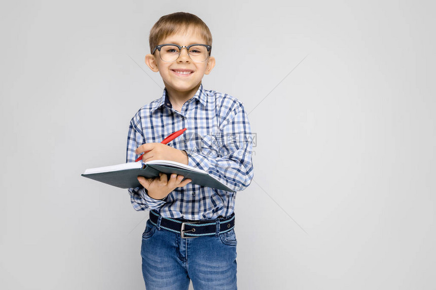 一个男孩在灰色背景上的肖像一个戴眼镜的男孩男孩手里拿着一本笔记本和一支红笔一个男孩在笔图片