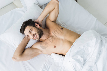 早上躺在白色床上微笑的赤膊男子的顶视图图片