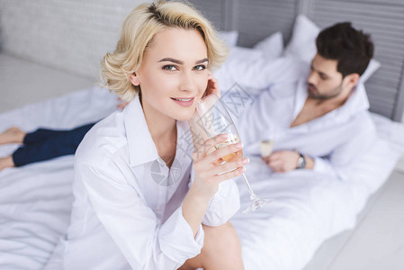穿着衬衫的年轻女子喝香槟看着镜头和男朋友在床上坐在图片