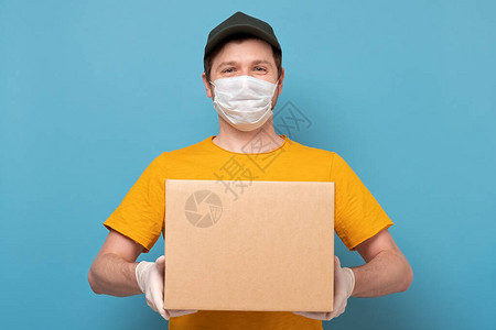 戴着医用面具的年轻送货员拿着一个蓝色背景上孤立的纸箱以检疫概念图片