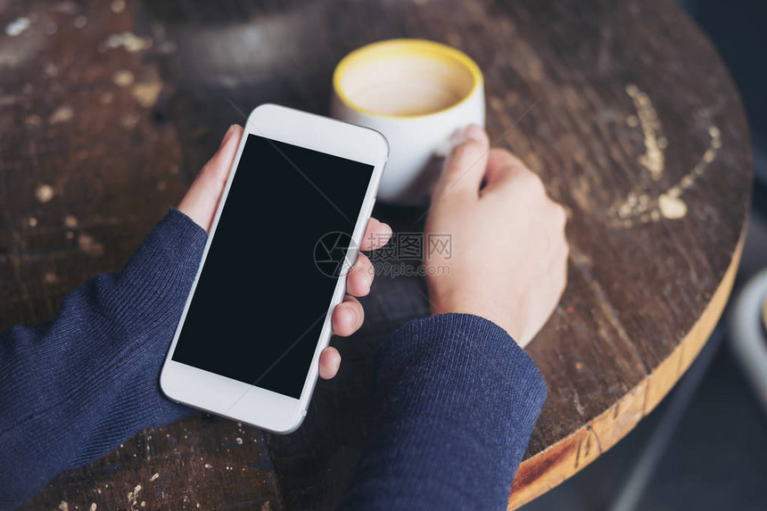 妇女手握白色手机空白黑屏和老咖啡厅木制桌上咖啡杯的混相图象图片