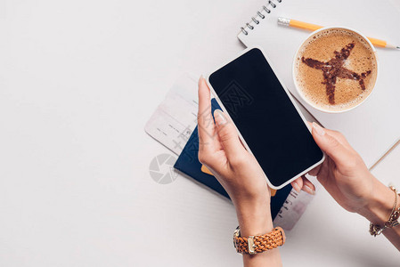 以笔记本咖啡杯和护照旅行概念在桌面上用空白屏幕显示手持智能手机的妇图片
