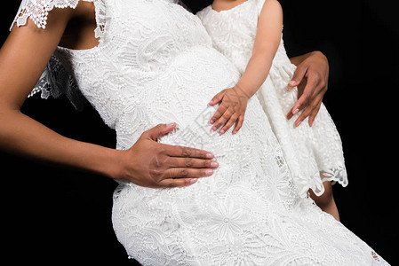 怀孕母亲和学龄前女儿触摸被黑孤立的腹部的图片