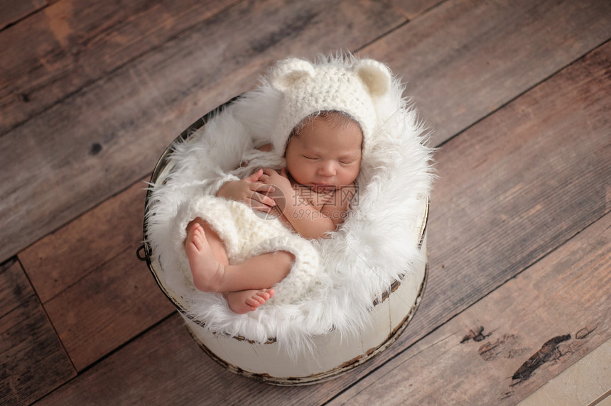 一个十一天大的新生女婴睡在木桶里的俯视图她戴着一顶钩针编织的白色小熊帽在仿古木背景图片
