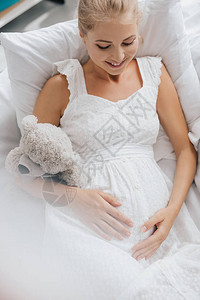 穿着白睡衣带着泰迪熊的白色睡衣微笑的孕妇在图片