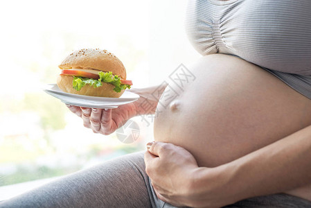 一个大肚子的孕妇在窗边拿着一个汉堡怀孕期间快餐和不健图片