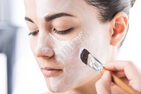 美容师在被白色隔离的女孩脸上涂抹奶油的裁剪图像图片