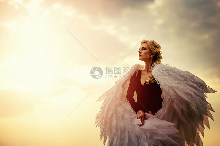 身着黑色礼服的年轻迷人女带着白色大翅膀在日落时空背景上闪发光图片