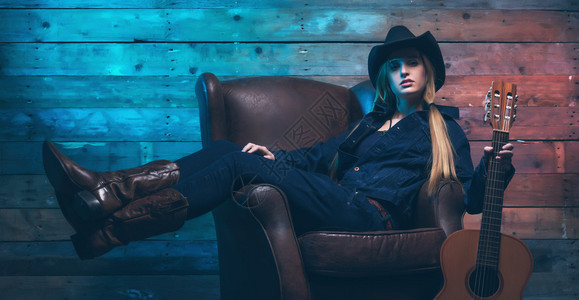 有原声吉他的女牛仔乡村歌手坐在皮椅上穿着蓝色牛仔裤和棕色图片