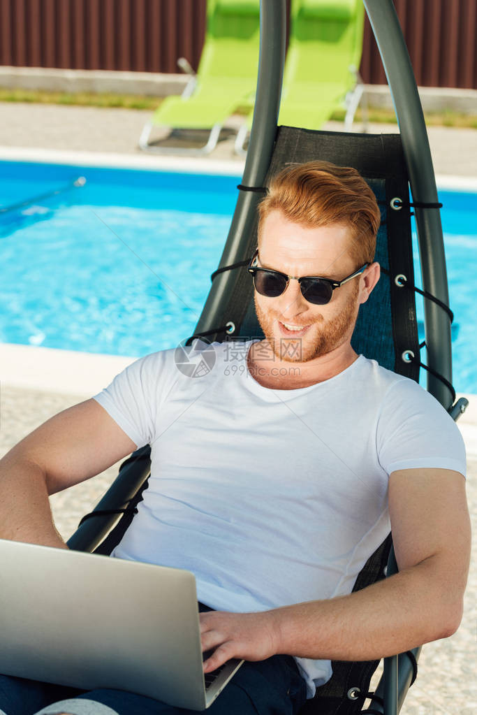 英俊的年轻人坐在游泳池前面的遮晒室用笔图片