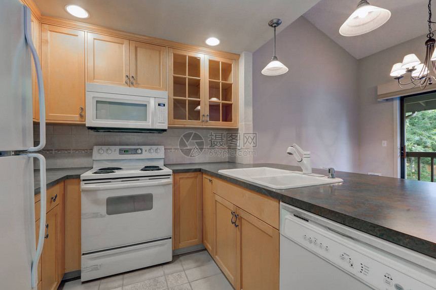 整洁紧凑的厨房配有枫木橱柜深灰色柜台白色电器和米色瓷砖后挡板图片