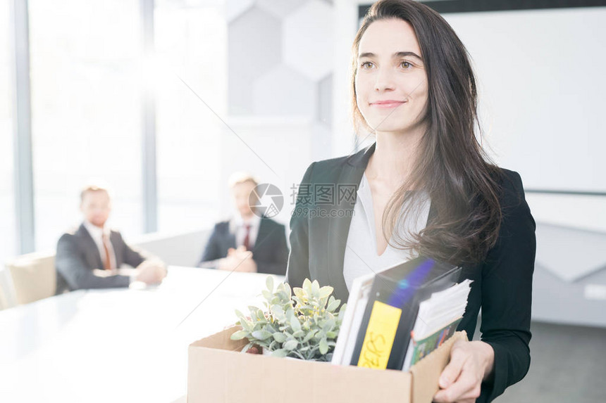 将微笑的年轻商业女商人在辞职后拿着一箱个人物品离开办公室的肖图片