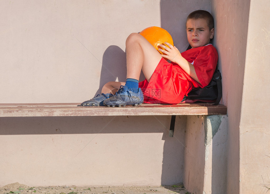 一个穿着红色足球衫的悲伤小男孩坐在长凳上图片