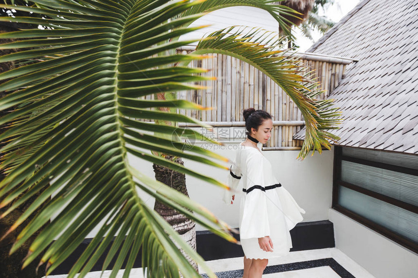 白色束腰外衣的女人在有棕榈树和白色背景的热带花园里在私人别墅拍摄时装图片