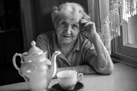 老年妇女坐在茶壶旁边的厨房桌前喝着图片