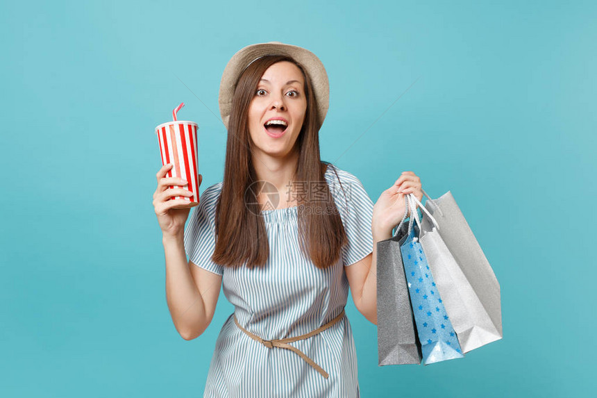 穿着暑期服装的漂亮时髦笑脸女孩拿着包装袋的草帽和购物后购买在蓝面糊背景上隔绝的苏打水杯图片