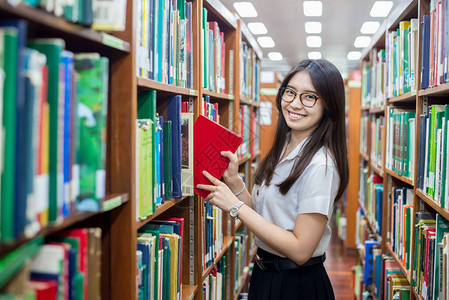 快乐的亚洲女学生在课间休息期间站在大学现代室内图书馆架附近的文学课上阅读背景图片