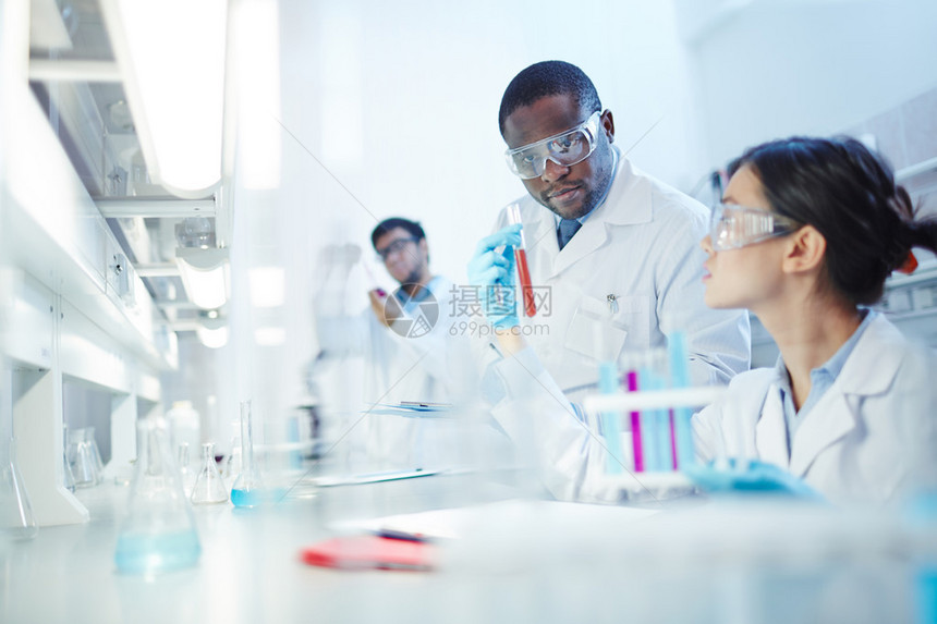 身着实验室外套和安全护目镜的亚洲女实验室科学家向实验室里好奇的非洲裔美国同事展示装有液体的试管背景中的拉丁美洲科学家在实图片