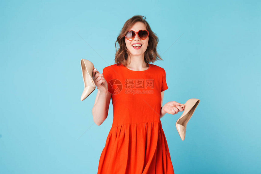 穿着衣服和心墨镜的年轻微笑的女士拿着经典鞋子站在蓝图片