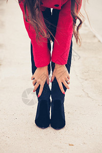 街上穿红色大衣的年轻女子触摸着她的黑色高跟鞋靴图片
