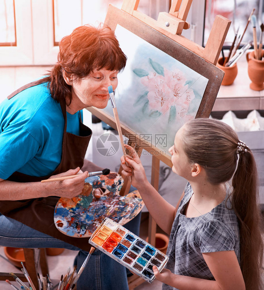 艺术家在工作室画架正宗的老妇人和孩子女孩用调色板水彩颜料调色板和刷子早晨的阳光私人企业绘图图片