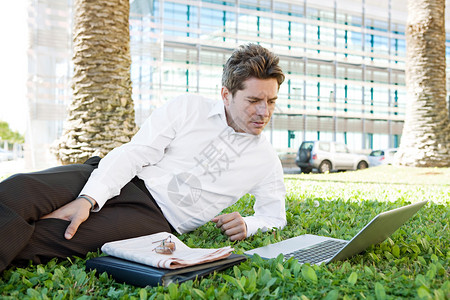 成功的商人在金融城区一栋现代玻璃办公楼附近的绿草上躺下图片