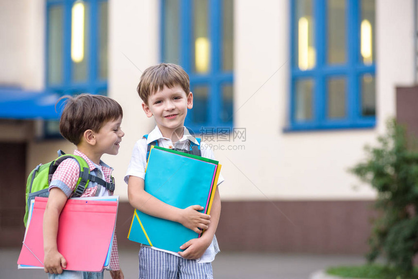 可爱的小学生在校园里轻快地交谈孩子们的心情很好温暖的春天早晨在同学的书包背后在其中一个男孩鲜红色图片