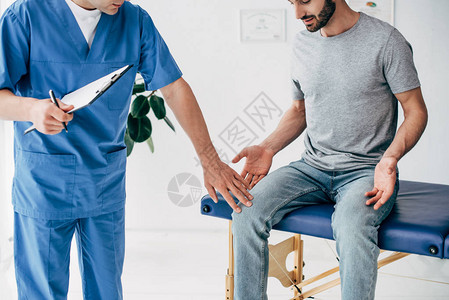 医生检查病人膝盖和把剪贴板放在诊所按摩柜里的切图片