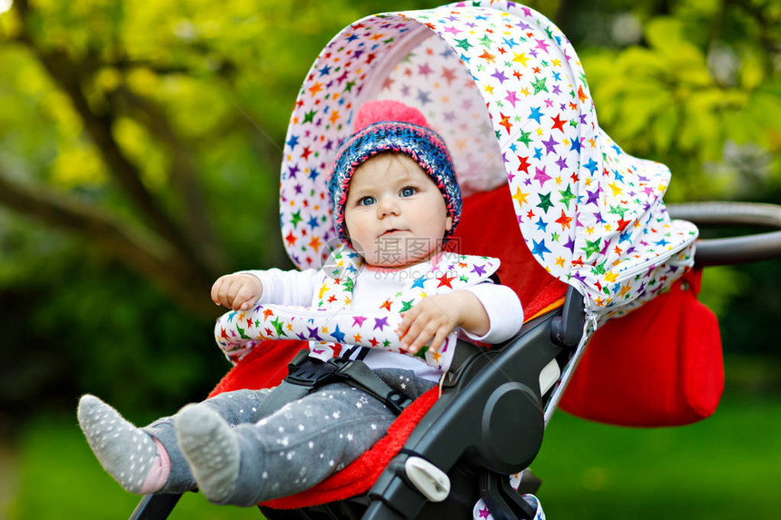 可爱的小漂亮女婴坐在时尚的婴儿车或婴儿车里等妈带着甜帽或子的快乐微笑的孩子有绿树背景宝图片