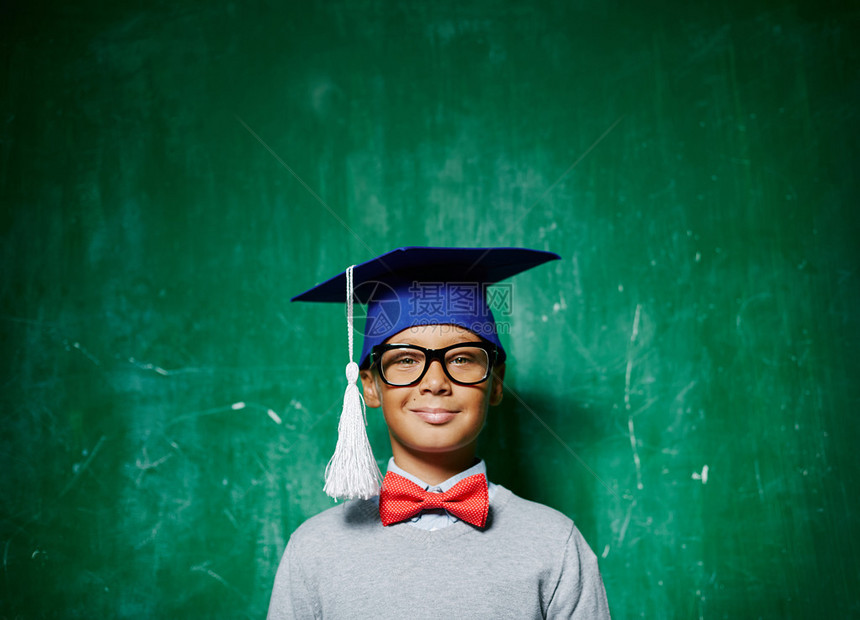 带着毕业帽子和眼镜微笑的男孩在看绿色黑板背图片